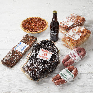Shipping packaging for Goode Co's The Independence meal bundle. Vacuum-sealed smoked brisket, pork ribs, link of Czech sausage, and link of jalapeño pork sausage. Glass bottle of Goode's Original BBQ sauce, and 2 loaves of jalapeño cheese bread.