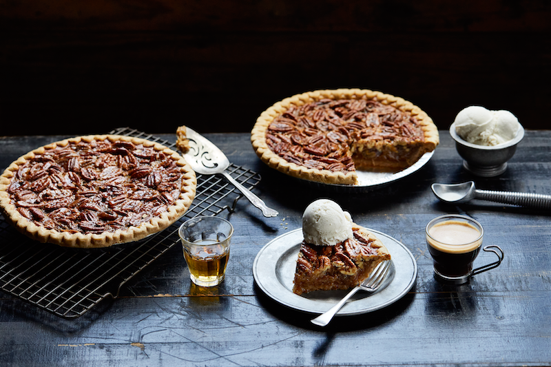 Two pecan pies, with a cup of coffee, a glass of bourbon, and a slice of one of the pies with ice cream on top.