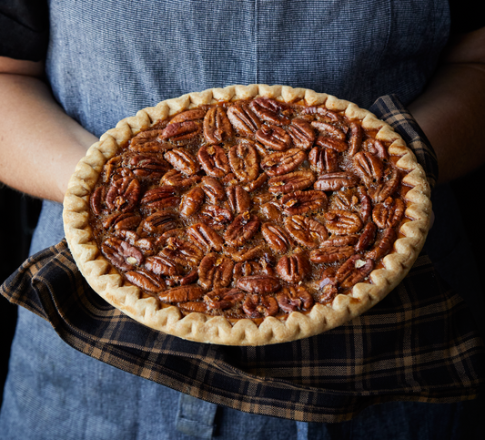 Person in a blue apron holding a Goode’s famous Brazos Bottom Pecan Pie in front of them, cradled by a blue and white towel.