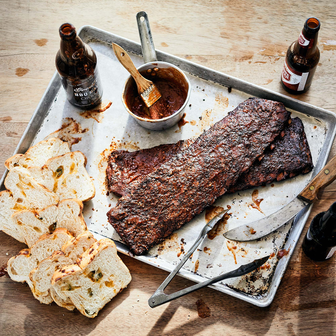 Two sets of ribs sitting on a metal cooking tray, next to sliced jalapeno cheese bread, tongs, and a bowl of Goode Co. BBQ sauce.