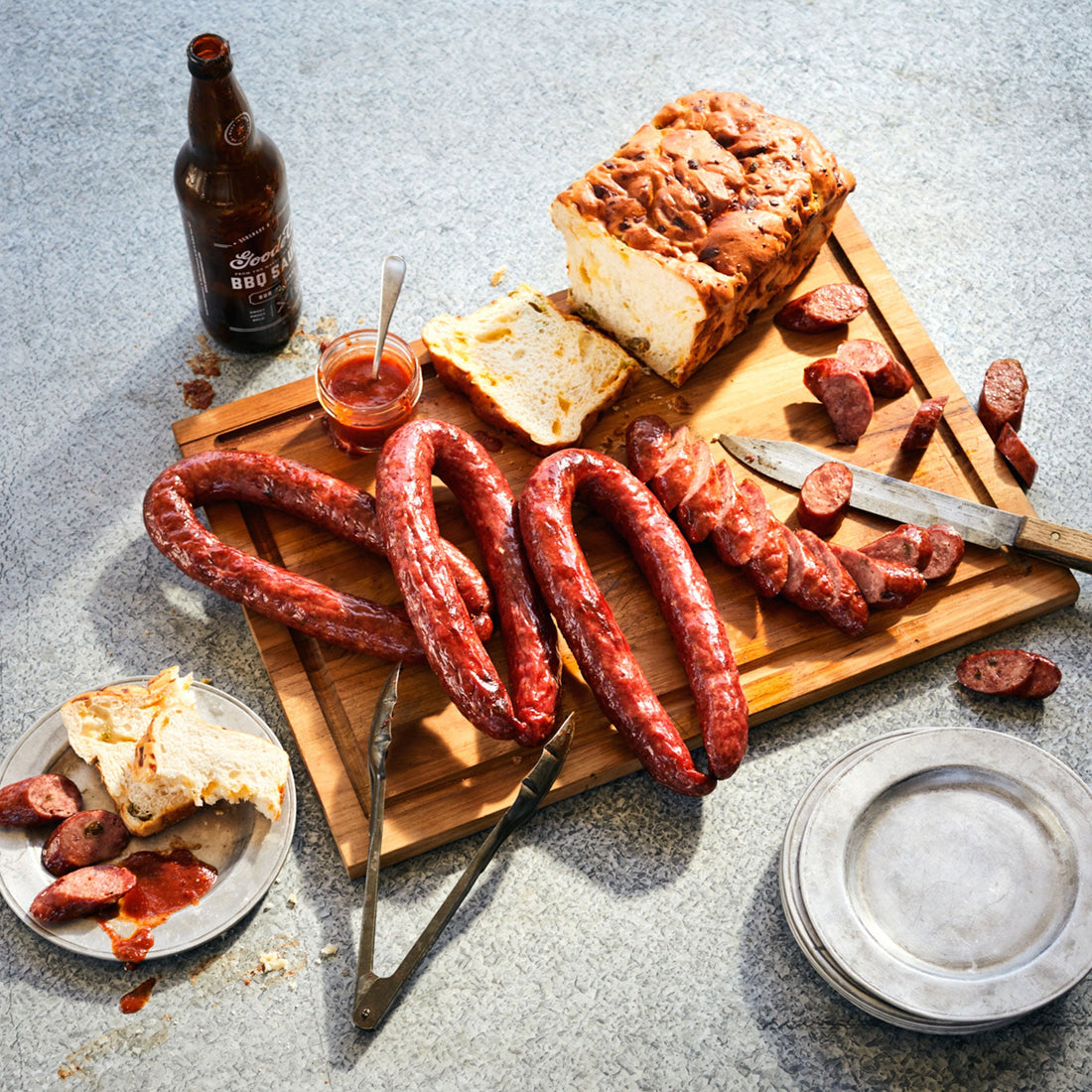 sausage sampler on a wooden cutting board with a loaf of sliced bread and a bottle of Goode Co. Sauce.