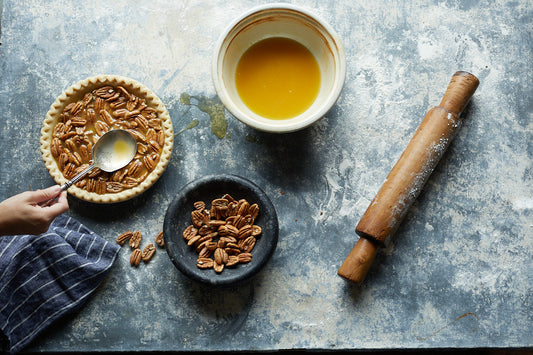 Flour-coated table with rolling pin, a bowl of pecans, melted butter, and an uncooked pie with a spoon hovering over it.