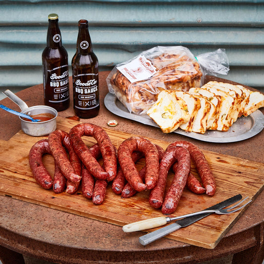 Uncooked sausage, two bottles of BBQ sauce, sliced, a package of a loaf of jalapeno cheese bread, and a cutting fork and knife.