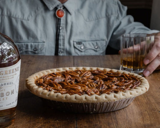 A pecan pie with a chocolate bourbon infusion, displayed on a table with a man in the background holding a glass of bourbon.
